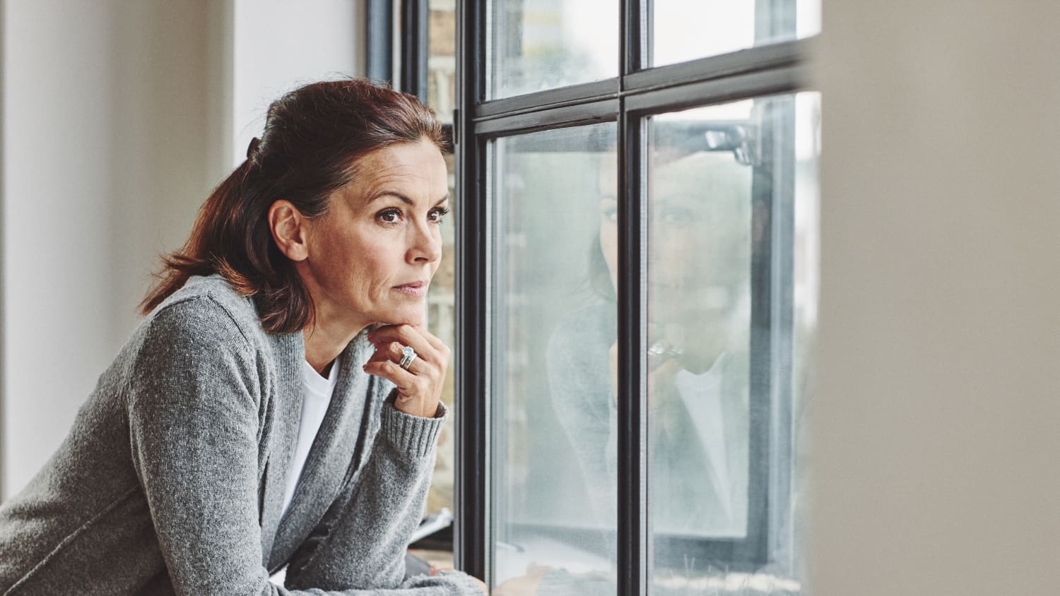 Thoughtful mature woman standing by the window, thinking about transvaginal mesh for pelvic organ prolapse