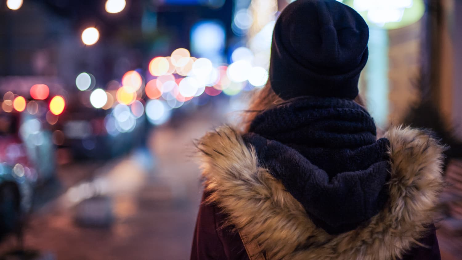 A teenager, who possibly has keratoconus, standing in a city at night, seen from the back of her head.