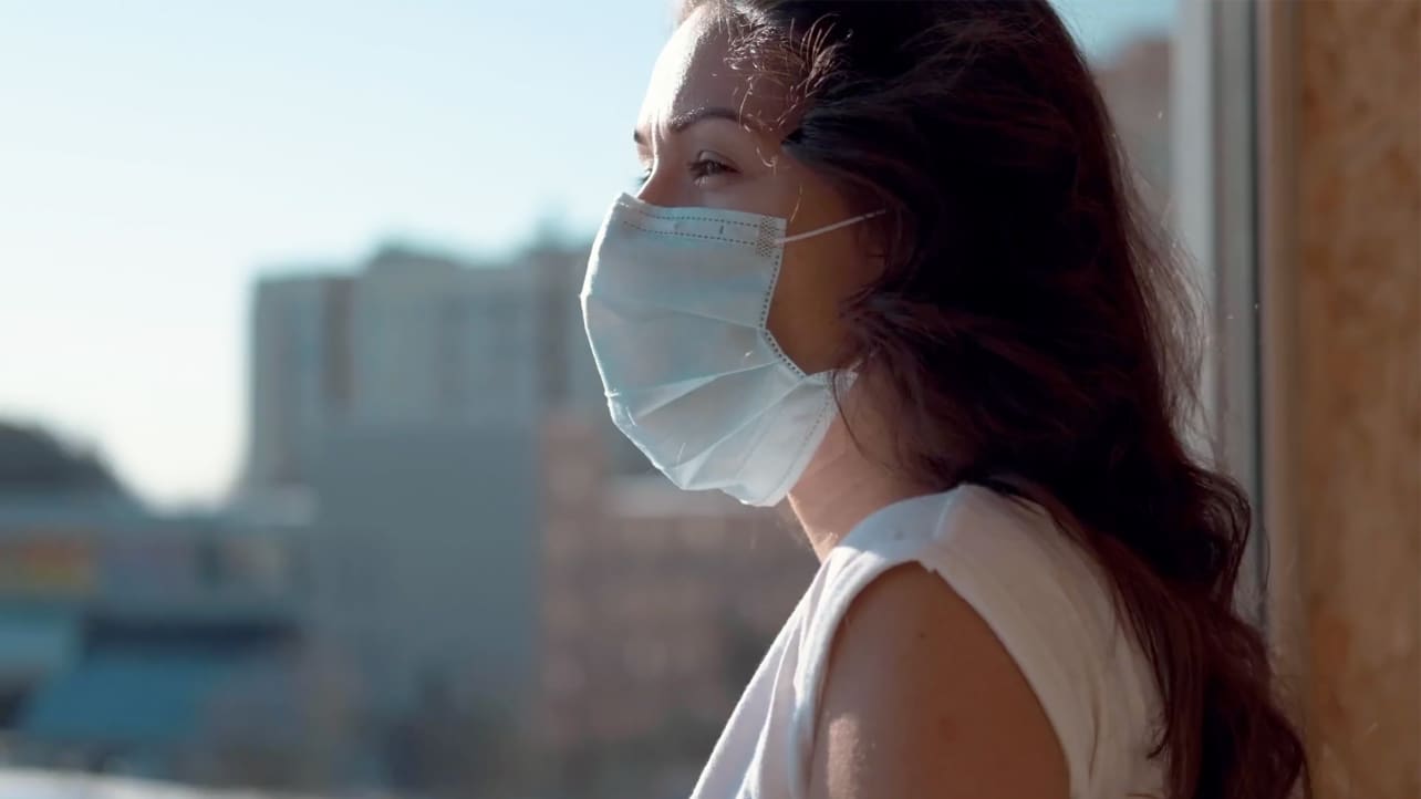 A woman wears a mask alone in her apartment during the COVID-19 pandemic.