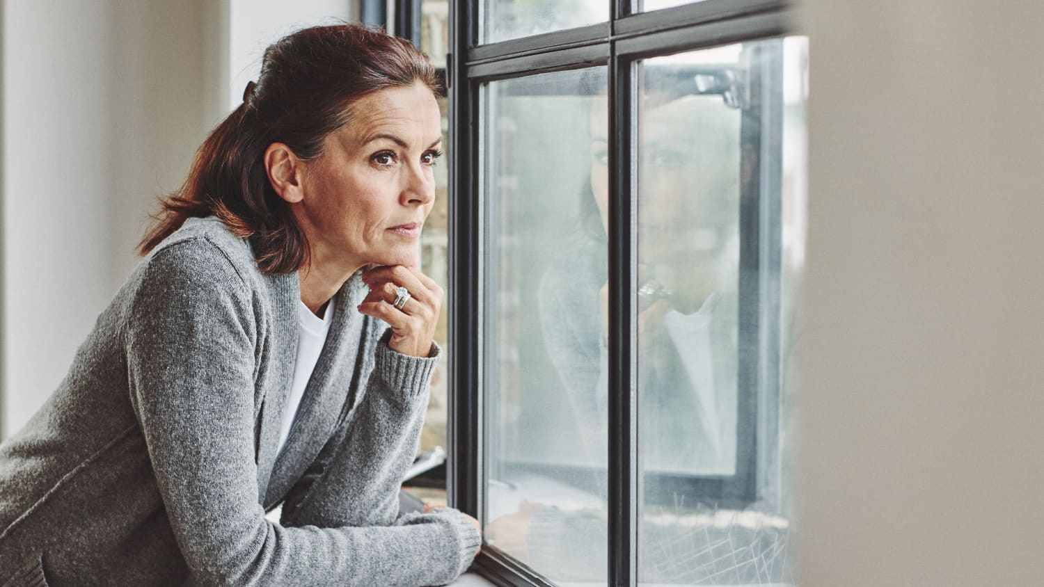 Thoughtful mature woman standing by the window, thinking about transvaginal mesh for pelvic organ prolapse