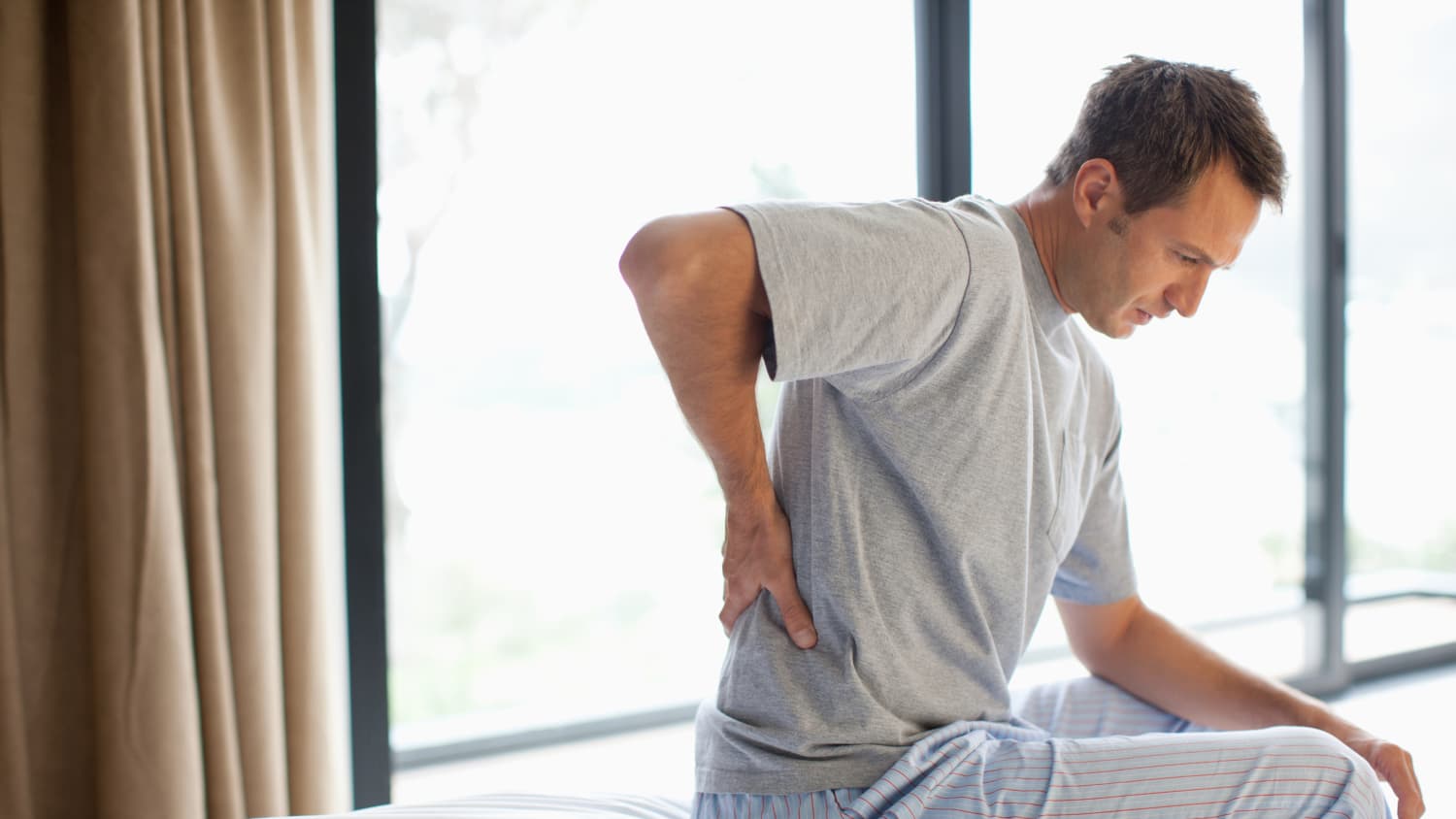 A man sits on his bed, rubbing his back, possibly from a herniated disc