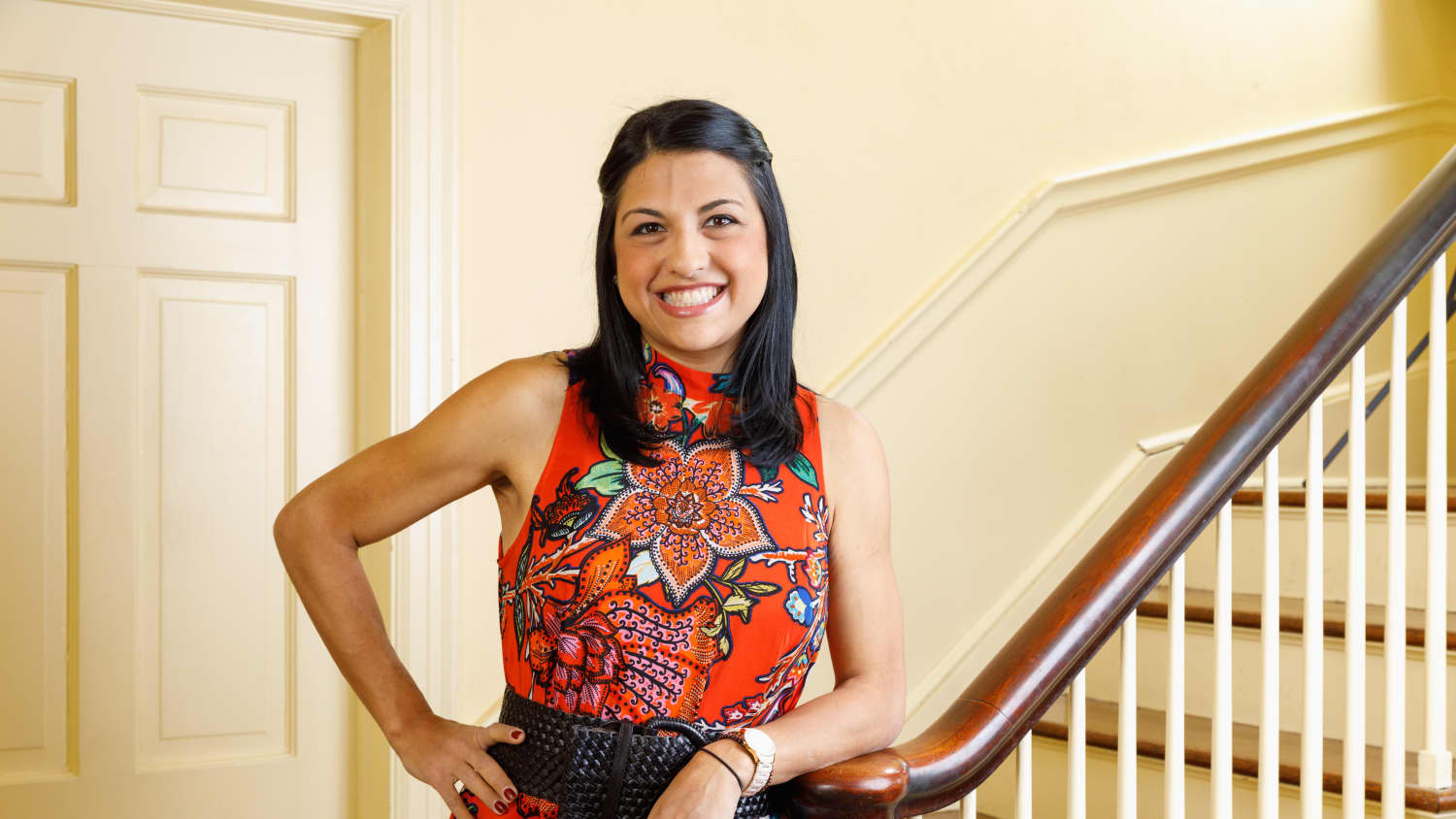 Amanda wearing a patterned red dress leans on a bannister. 