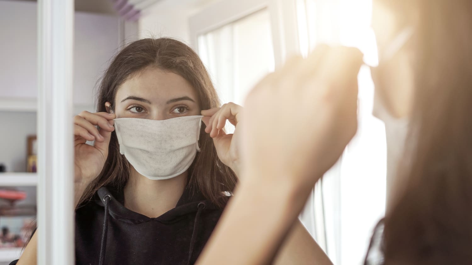 woman putting on a mask, possibly hiding maskne, in front of a mirror