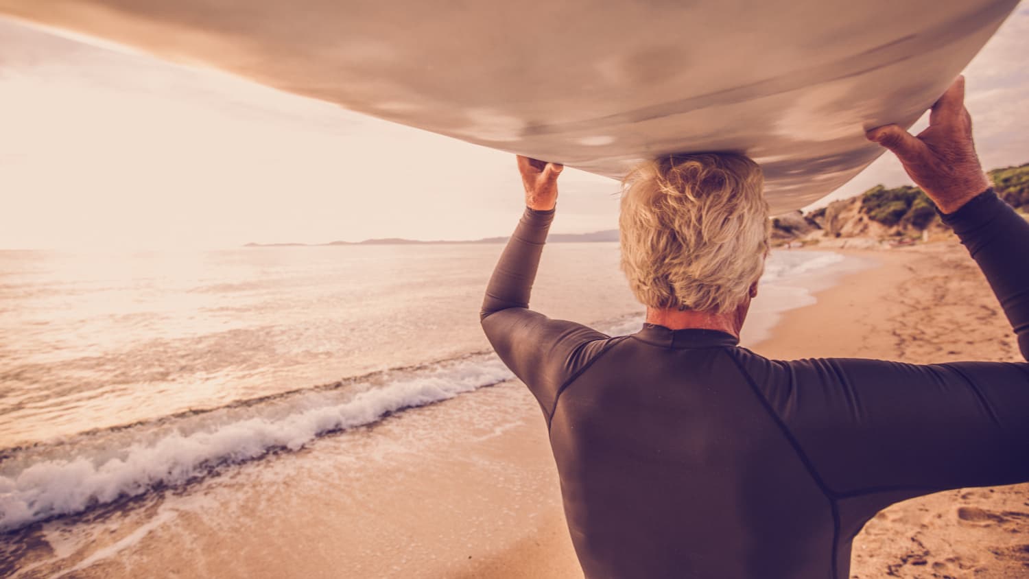 An older man, who possibly has Peyronie's disease, is shown carrying a surfboard along a beach.