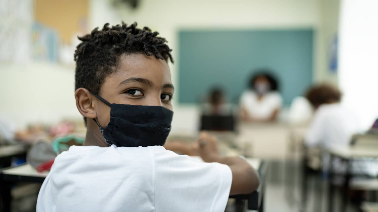 child in face mask in classroom, stressing the importance of COVID-19 vaccines