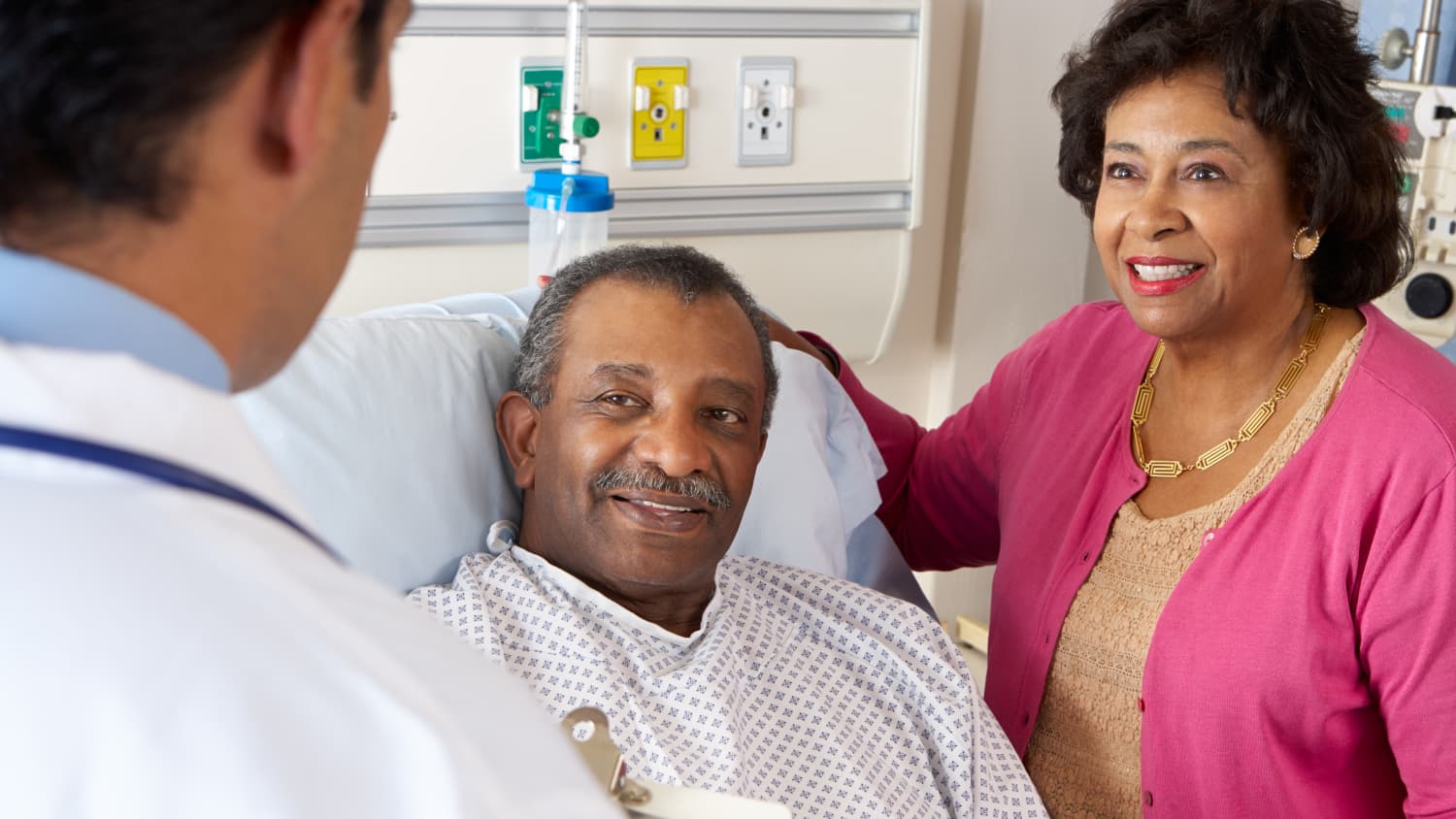 A man in a hospital bed talks to his wife (in a bright pink sweater) and his doctor after minimally invasive gastrointestinal surgery.