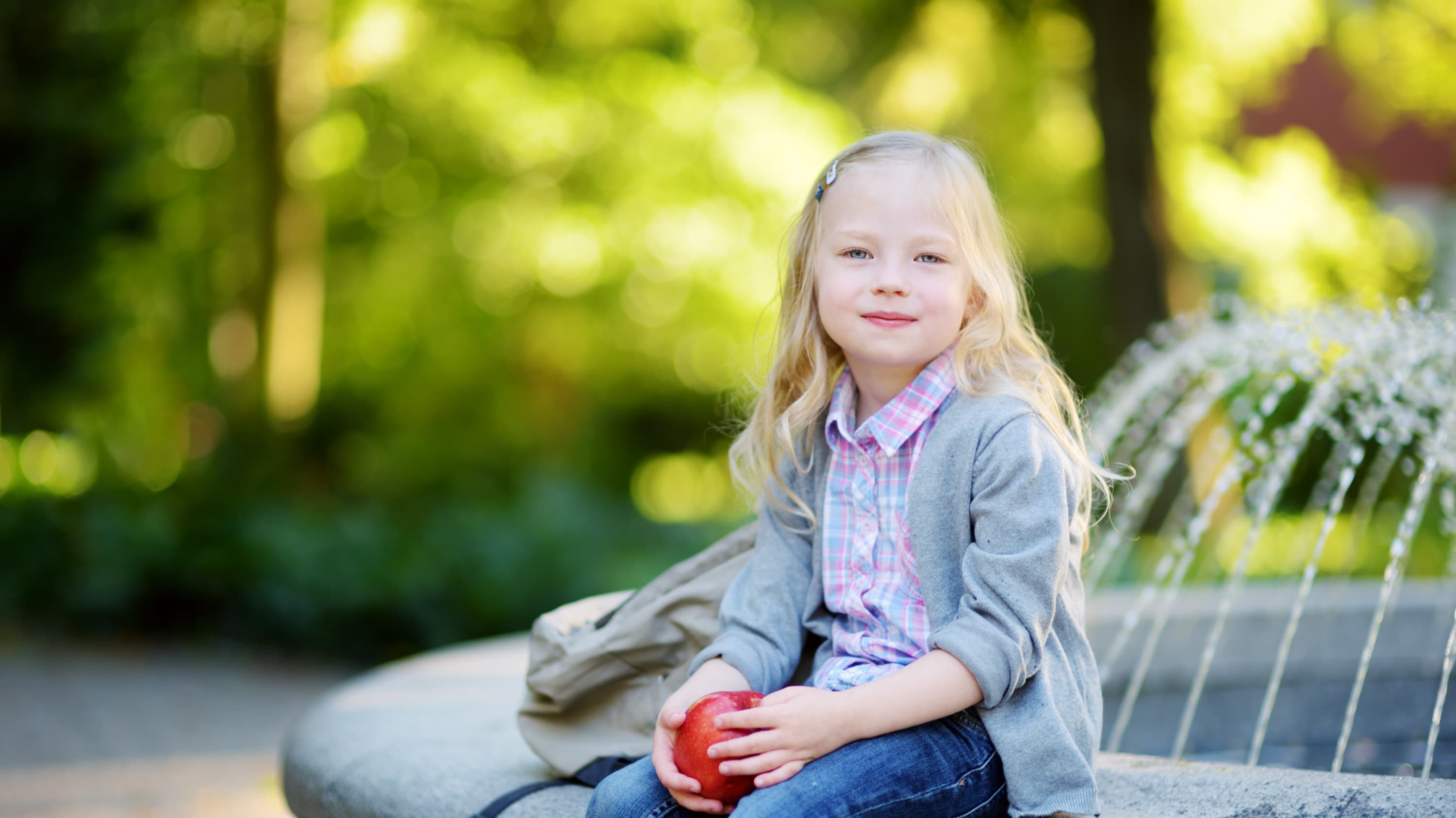 A girl, who could be a pediatric cancer survivor, is sitting on a the edge of a park fountain with an apple in her hand.