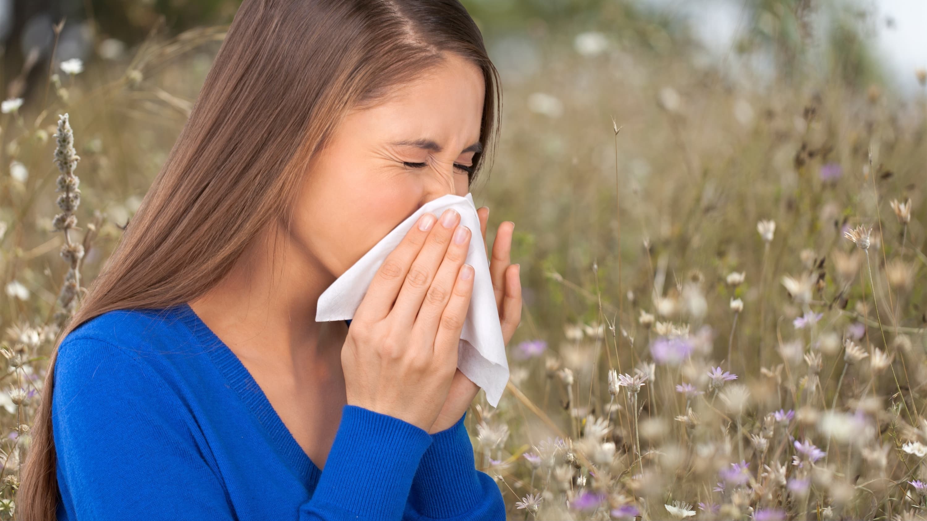 A woman in a blue shirt who has seasonal allergies sneezes into a tissue outside.