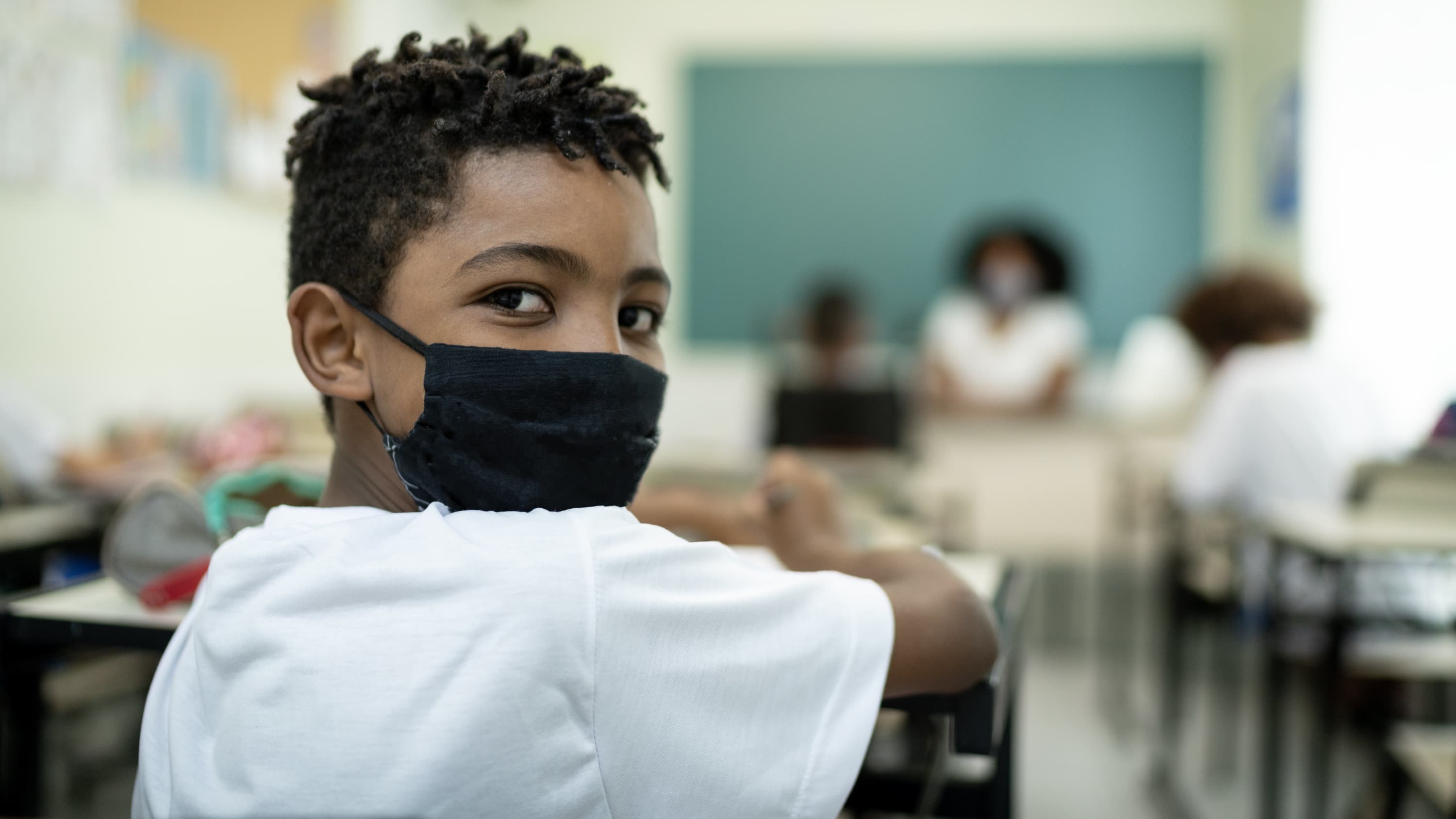 child in face mask in classroom, stressing the importance of COVID-19 vaccines
