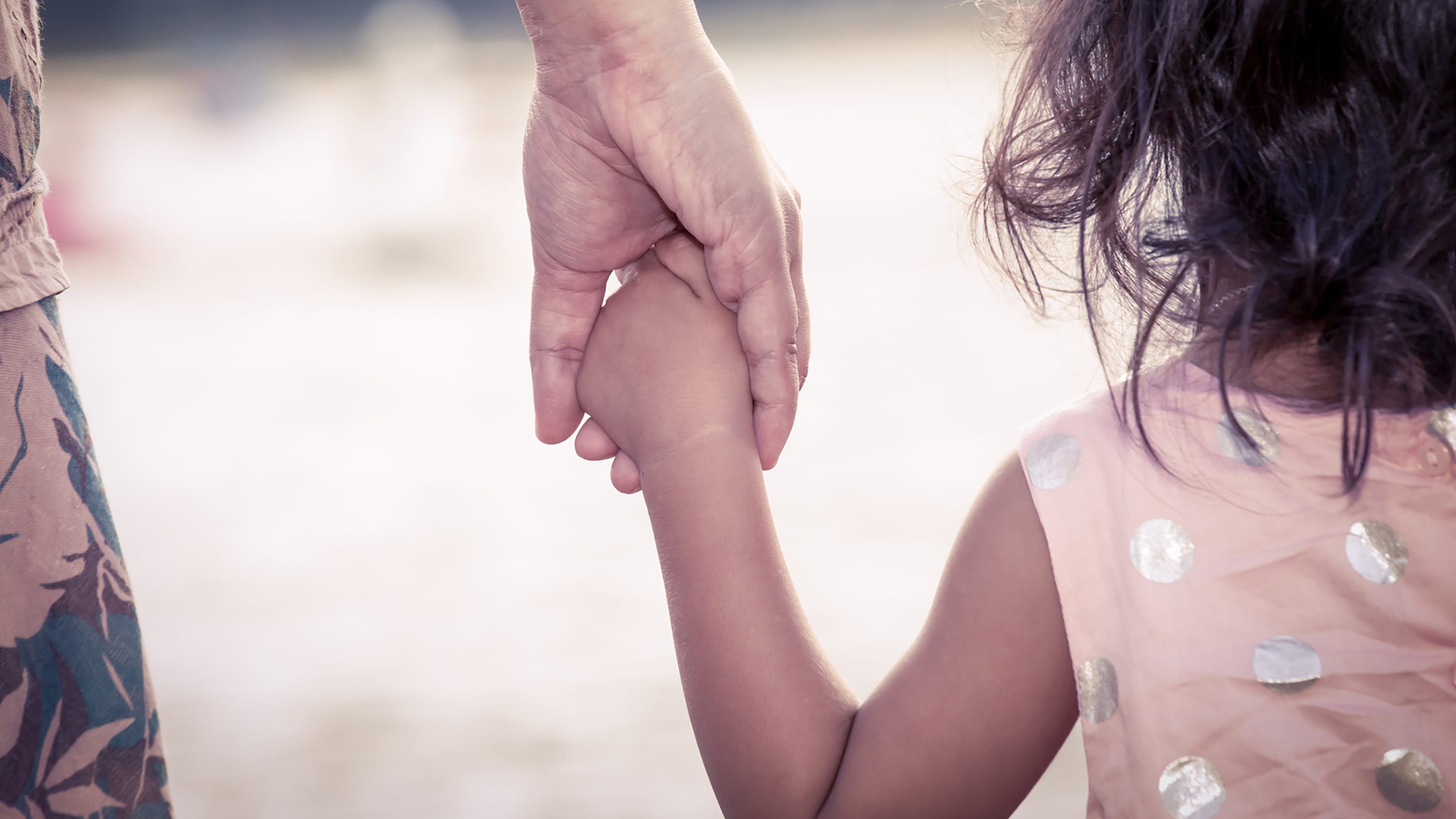 Little girl in a pink and white polka dot dress holds the hand of a parent who may be depressed.