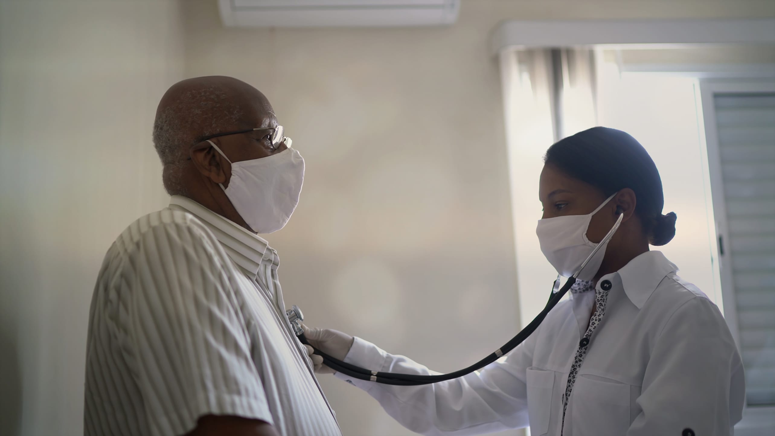 A masked doctor listens to a patient's heart.