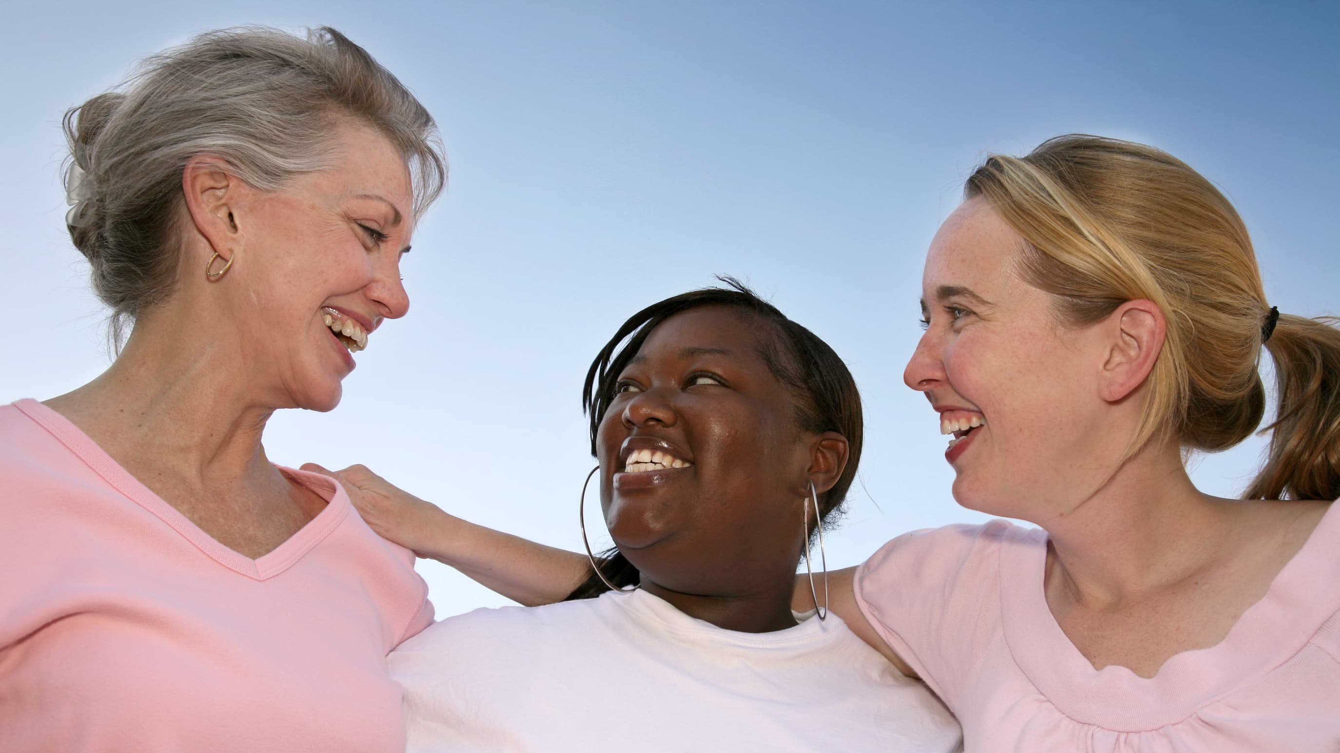 Three smiling women who could be cancer survivors lock arms.