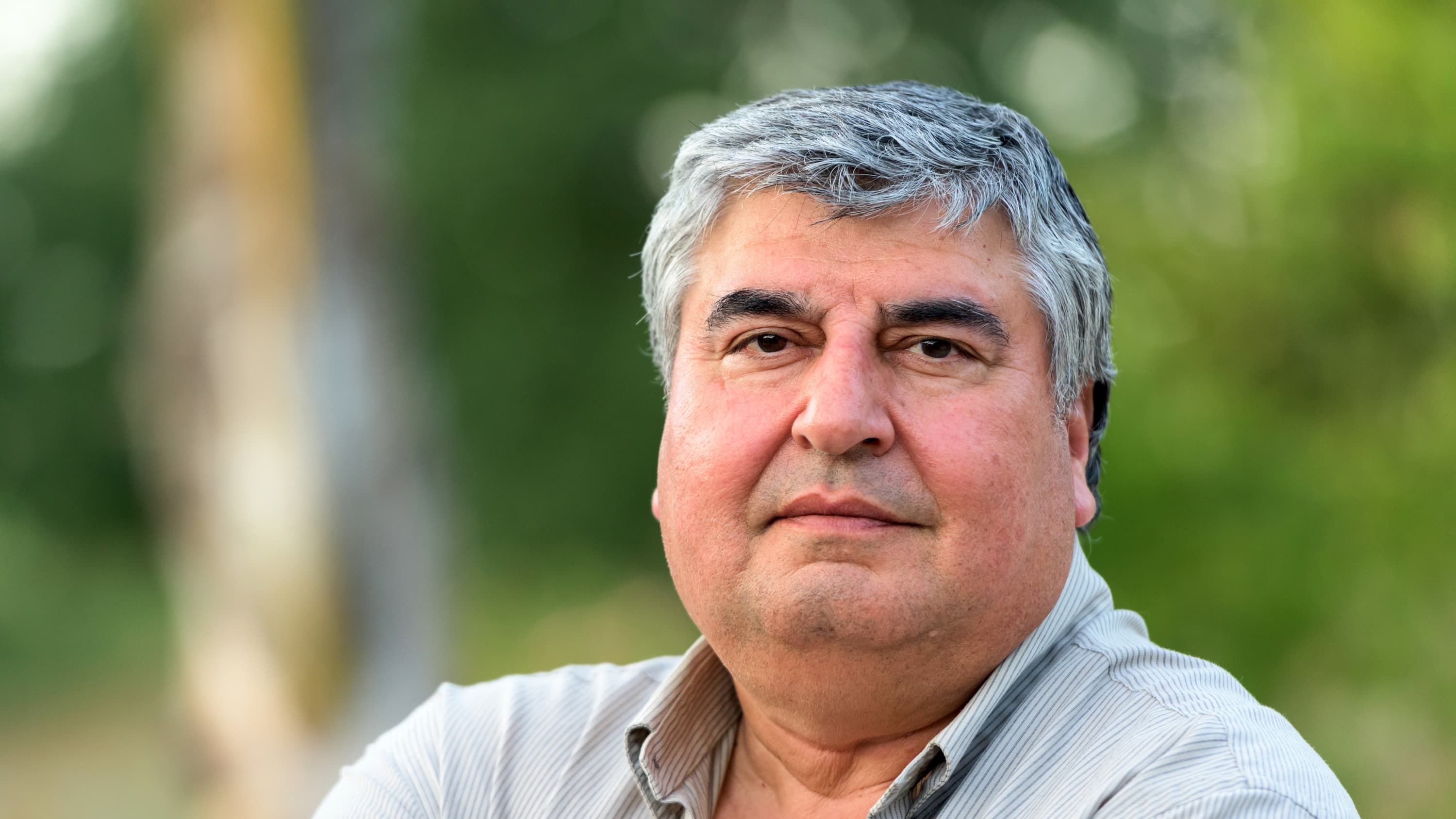 An overweight man who may have peripheral artery disease looks into the camera. He is wearing a grey polo shirt against a green, leafy background.