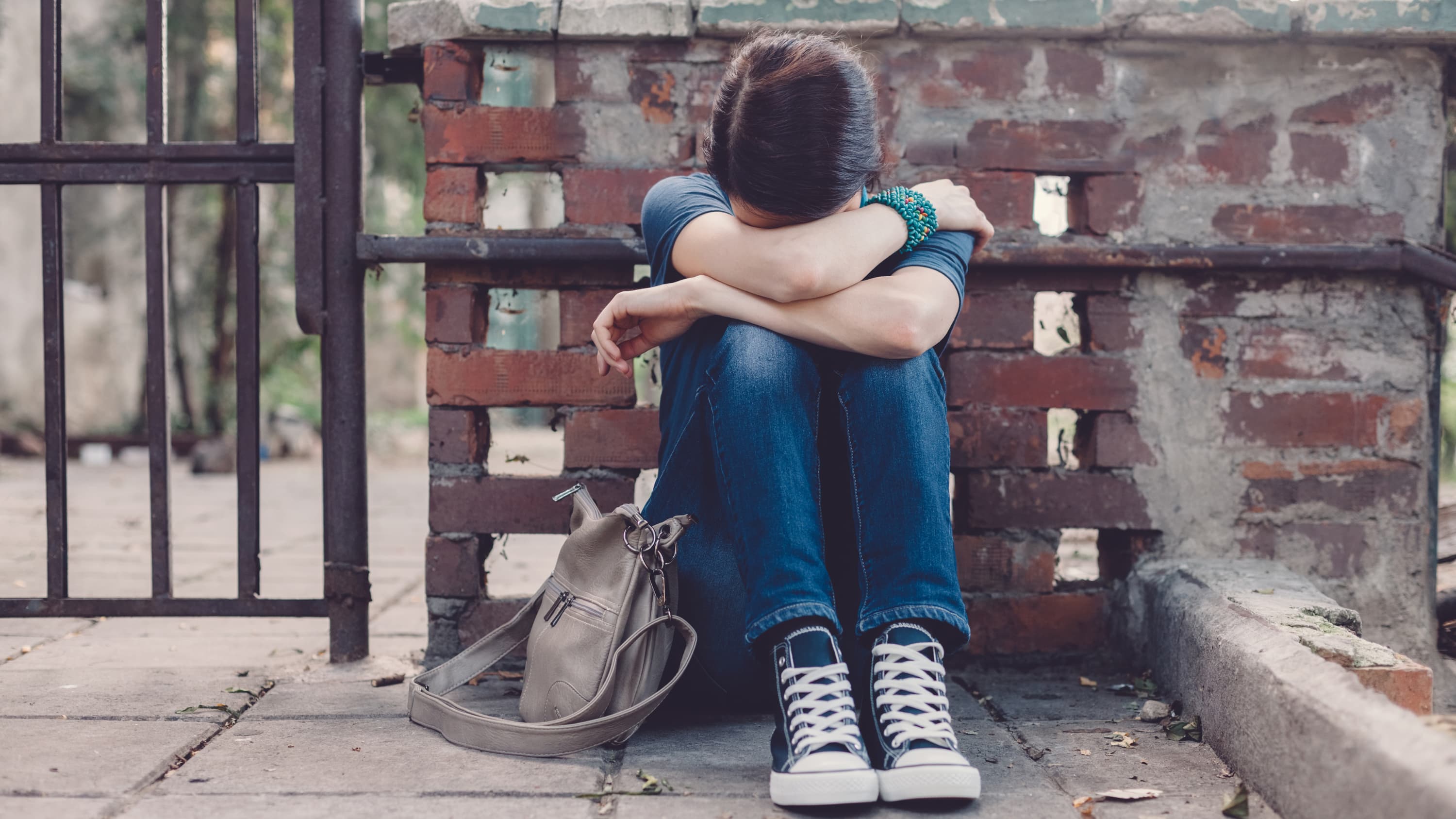 A boy who looks like he is feeling depressed sits on the ground beside his backpack with his head in his hands.