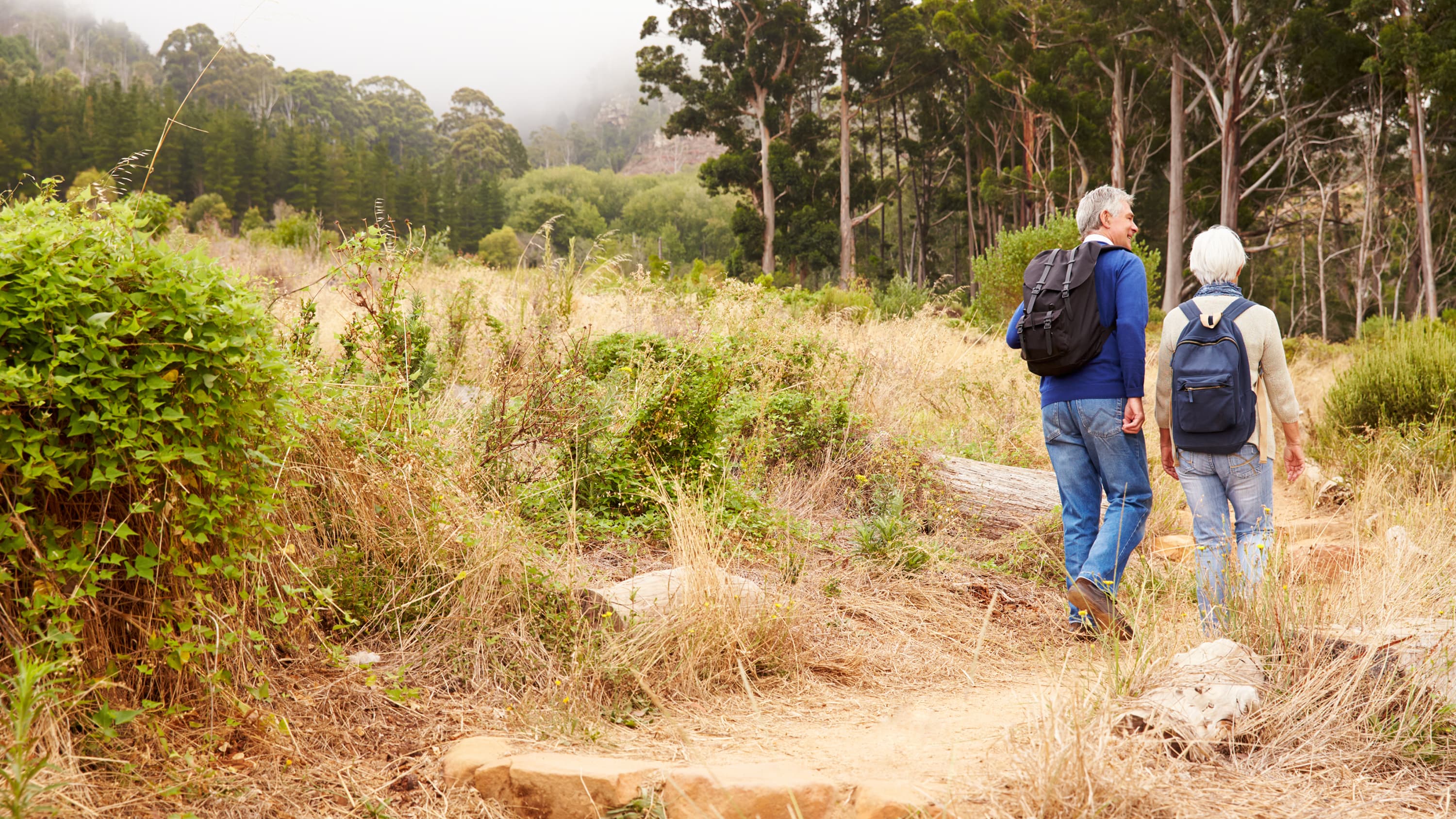 An older couple, one of whom has a spinal deformity, walk on a trail.