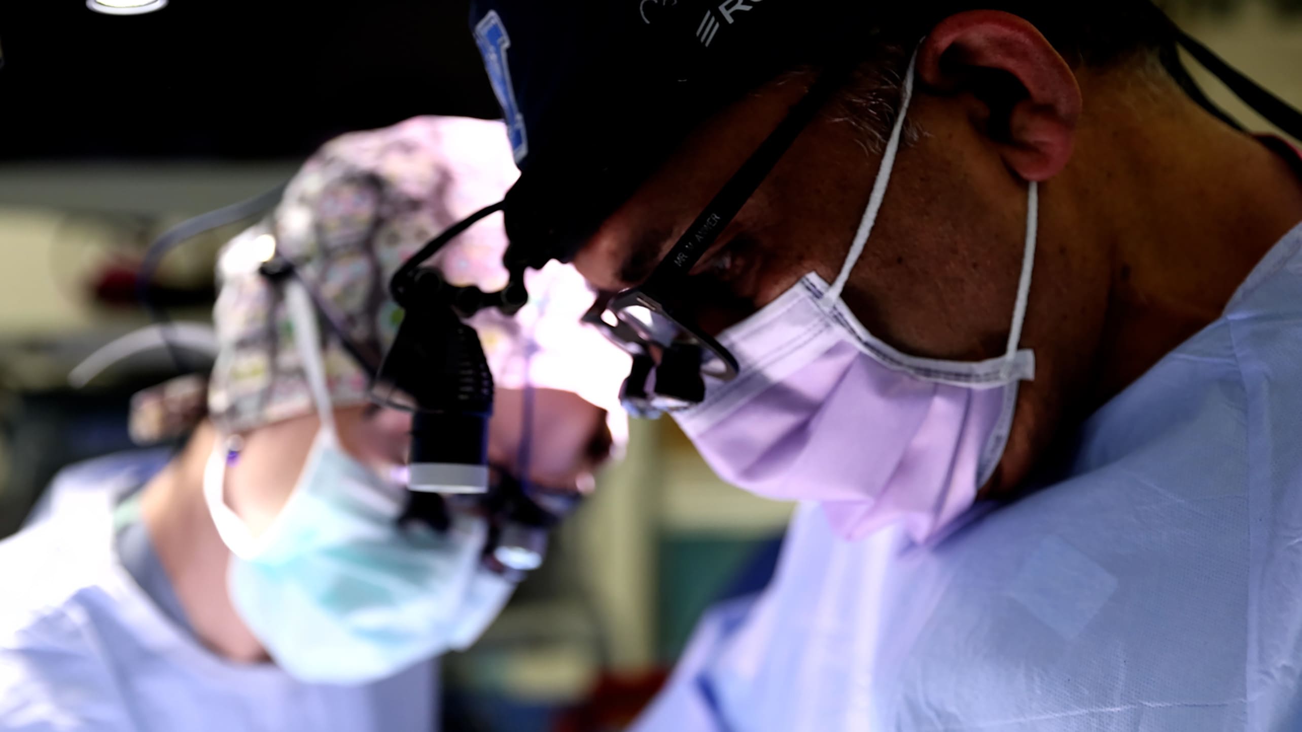 A closeup shot of a heart surgeon wearing surgical mask, scrub cap, and headlight looking down intently