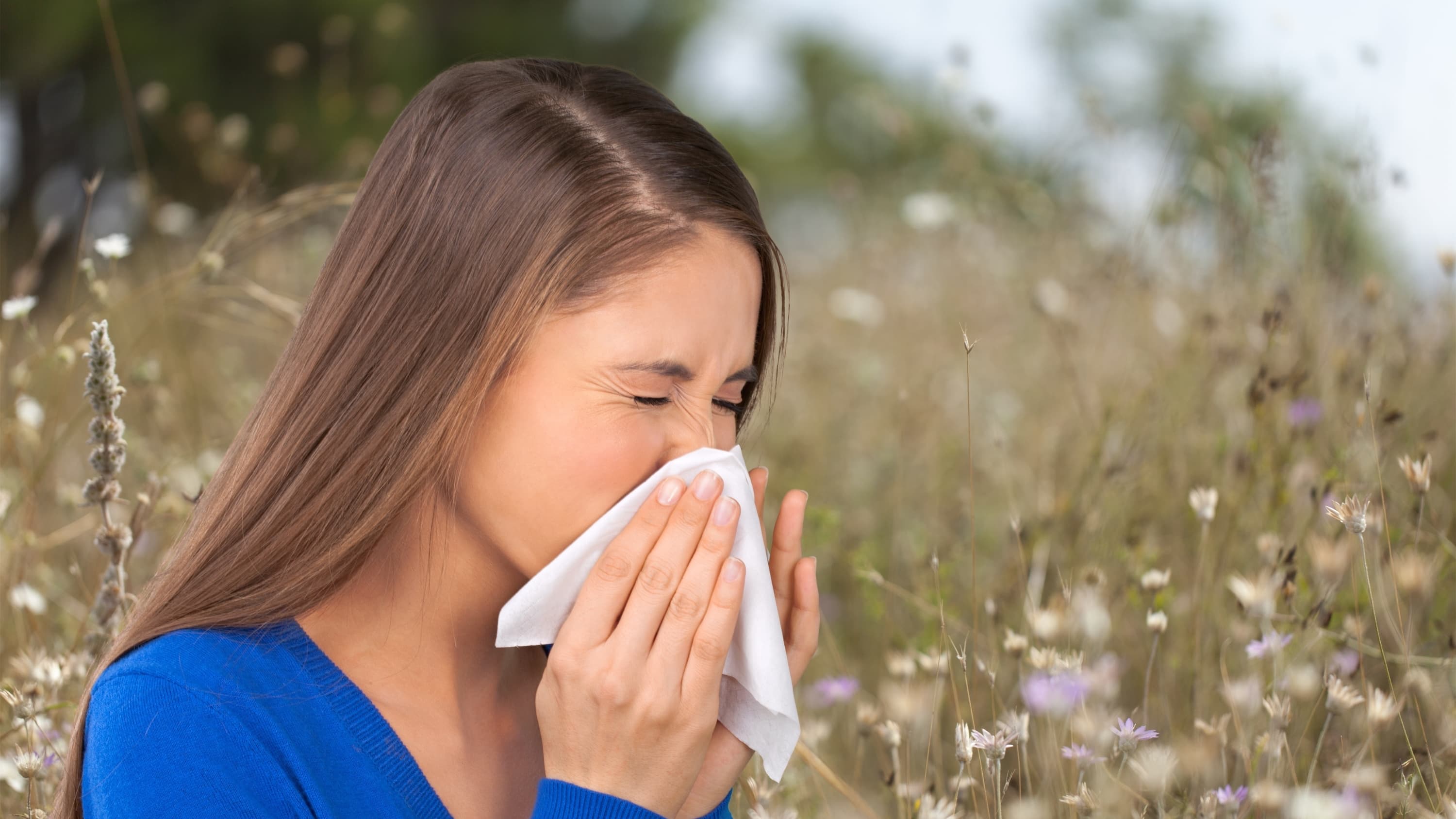 A woman in a blue shirt who has seasonal allergies sneezes into a tissue outside.
