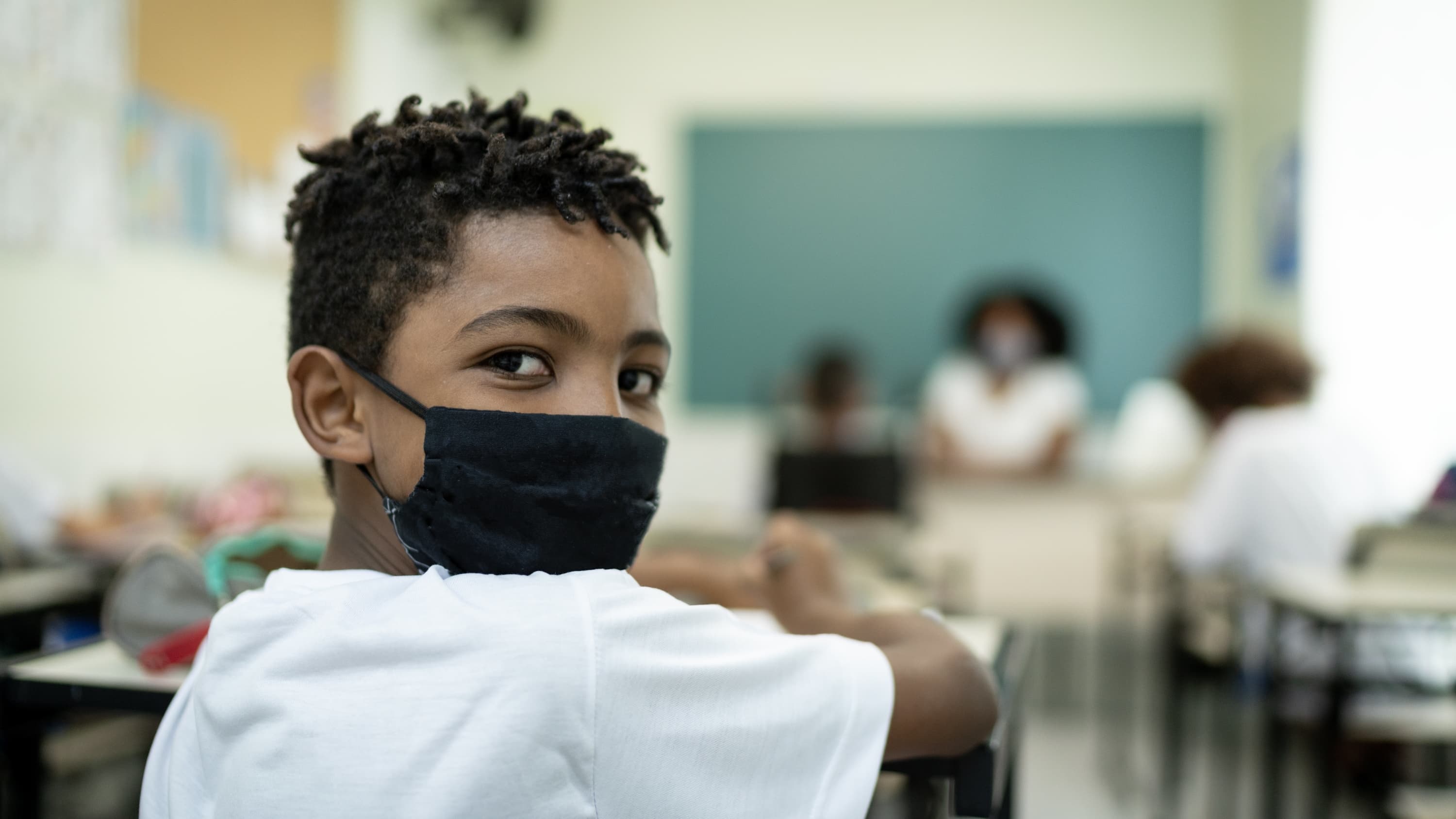 child in face mask in classroom, stressing the importance of COVID-19 vaccines
