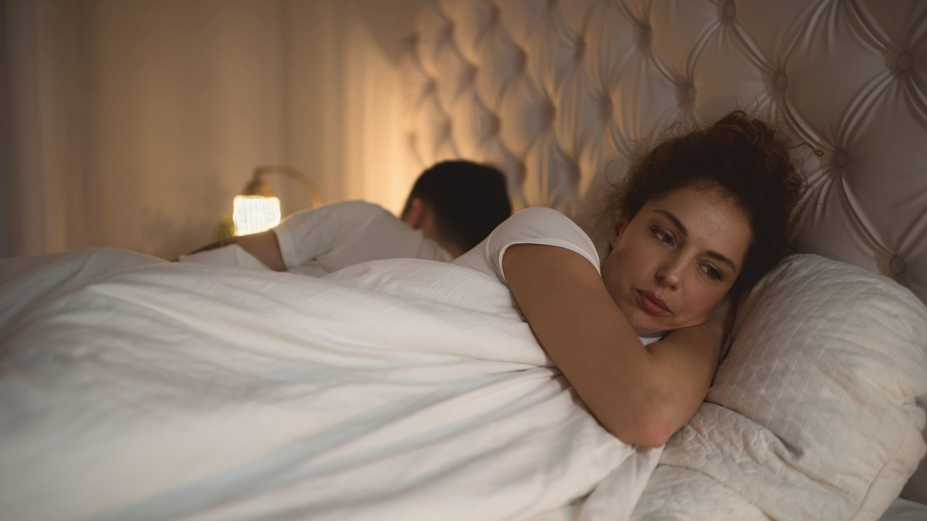 Woman lying in bed awake with her partner next to her asleep.
