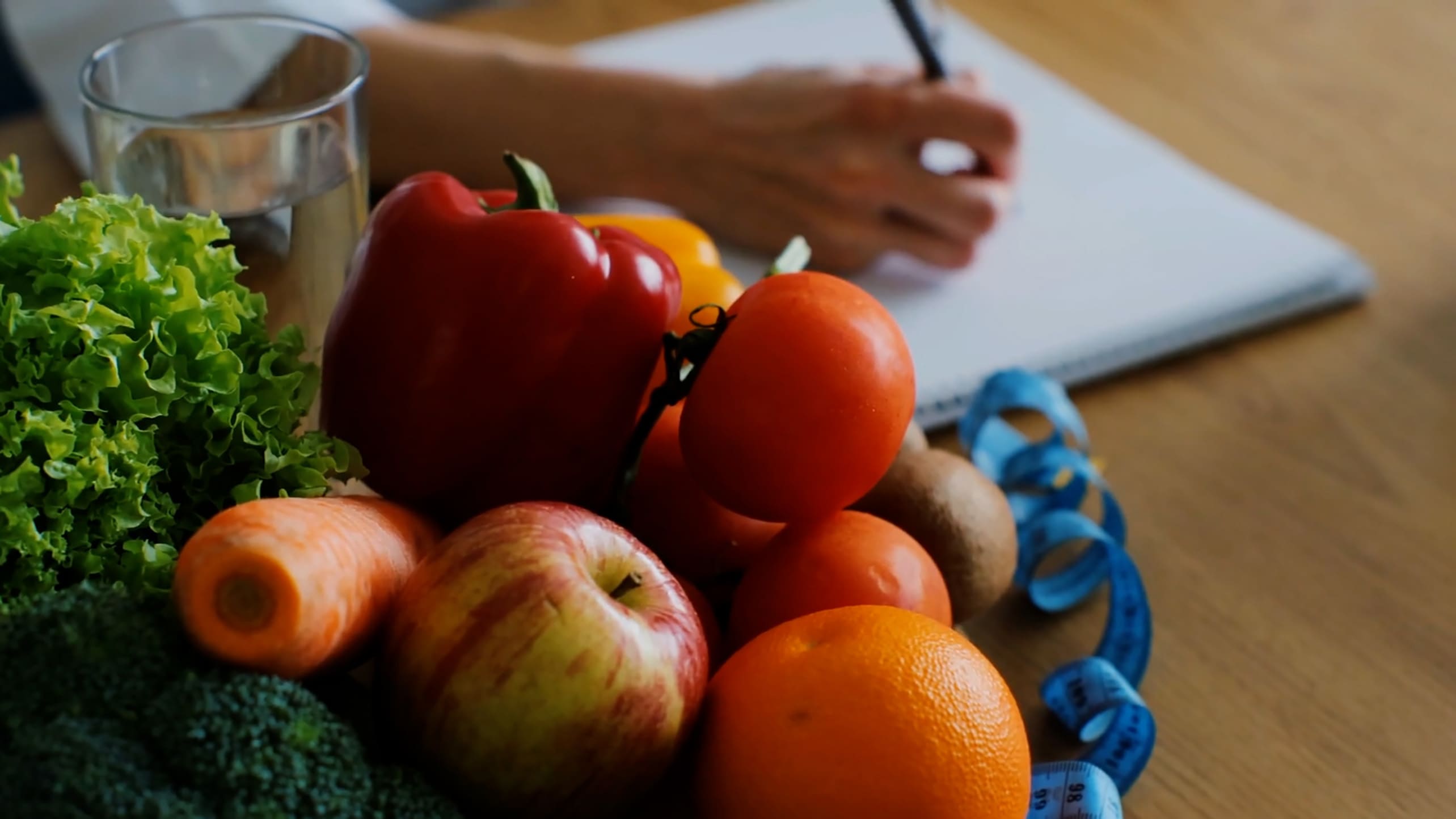 A small pile of fresh fruits and vegetables in the foreground with a hand holding a pen and writing in a notebook in the background