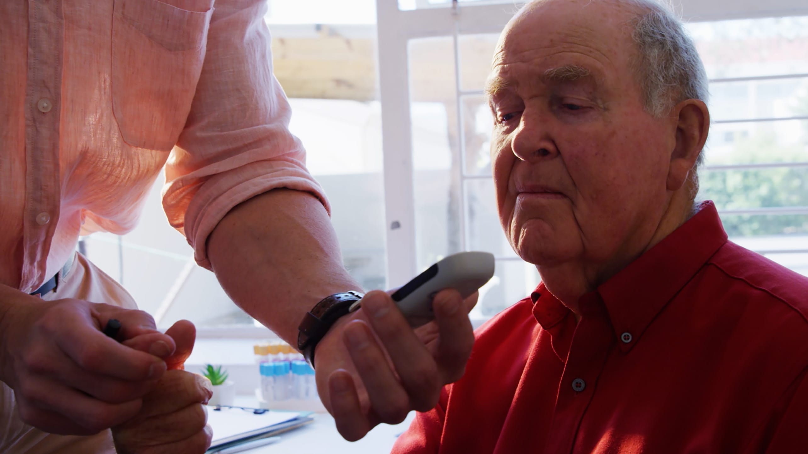 An elderly white man looks at a glucometer held by another person's extended hand